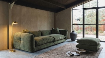 Un sofá de pana verde oliva ocupa una sala moderna con paredes texturizadas, una lámpara dorada, una ventana panorámica y una alfombra marrón con un libro.