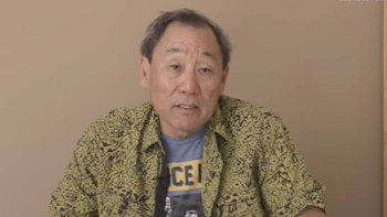 Primer plano de un hombre mayor con pelo canoso y piel clara, vestido con una camisa verde estampada sobre una camiseta azul, mirando al frente