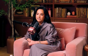 Becky G sentada en un sofá rosa pálido, vestida con una camisa y pantalones de rayas grises. Hay un micrófono frente a ella y una estantería de libros detrás