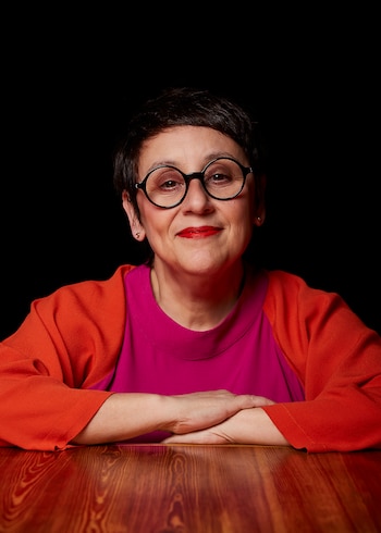 Retrato de una mujer sonriente con pelo corto oscuro y gafas redondas negras, vestida con una blusa fucsia y naranja, con las manos entrelazadas sobre una mesa de madera