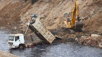 Un camión volquete sumergido en un río descarga material mientras una excavadora amarilla se encuentra en la orilla junto a tierra y escombros