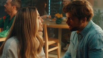 Una mujer de perfil y un hombre con barba sentados frente a frente en un salón con mesas y otras personas de fondo