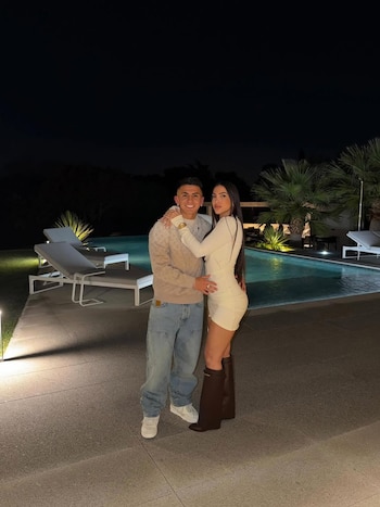 Thiago Almada y Antonella D'Alotta de pie, abrazados, junto a una piscina exterior iluminada por la noche con palmeras de fondo