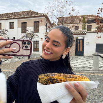 Mujer sonriente tomando una foto con un teléfono inteligente, sosteniendo un maíz asado. Fondo con edificios blancos y árboles sin hojas