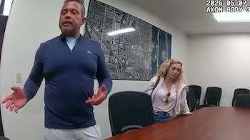 Luis Salazar, un hombre de cabello gris en camisa azul, gesticula ante una mesa. Una mujer rubia se apoya en la mesa detrás de él. Hay un mapa en la pared