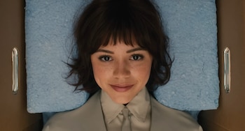 Mujer con cabello oscuro y flequillo, con pecas, acostada sobre un acolchado azul, dentro de una estructura marrón con dos asas metálicas
