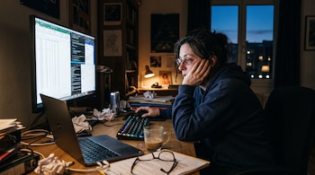 Una mujer apoyada en su mano observa la pantalla de una computadora junto a una taza de café y papeles arrugados.