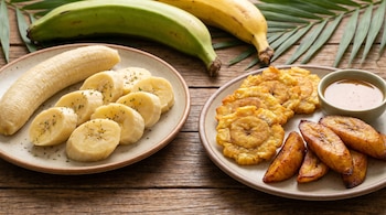 Dos platos de plátano macho sobre una mesa de madera. Un plato contiene plátano cocido en rodajas y entero, el otro tostones, plátano maduro frito y una salsa.