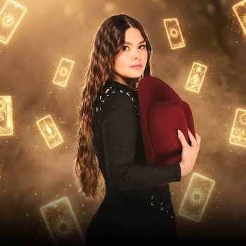 Yuridia sostiene un sombrero rojo vestida de negro, rodeada por ilustraciones iluminadas de cartas de tarot en el fondo