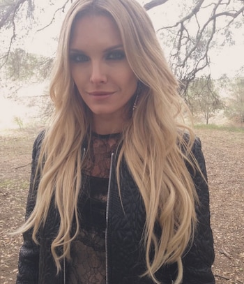 Mujer rubia de cabello largo y ondulado con un top de encaje negro y chaqueta acolchada negra, posando al aire libre con árboles de fondo
