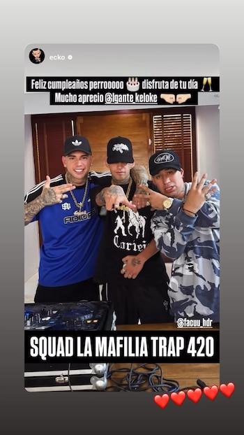 Tres hombres, incluyendo a L-Gante en el centro, posan sonrientes y con tatuajes visibles, haciendo gestos con las manos frente a equipo de DJ y puertas de madera