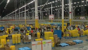 Interior de un amplio centro de distribución de Amazon con trabajadores, cajas, palés y equipos operando bajo un techo alto con iluminación brillante
