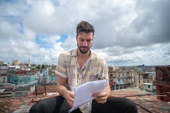 Hombre barbudo sentado en una azotea, sosteniendo y mirando papeles. Viste camisa estampada. Al fondo, edificios de ciudad bajo cielo nublado