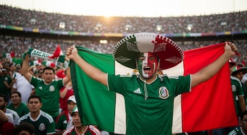 Hombre con sombrero de charro y cara pintada con los colores de México sostiene una bandera mexicana, gritando en un estadio lleno de aficionados.