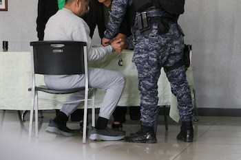 Un hombre con ropa deportiva gris sentado en una silla mientras un agente de uniforme camuflado le sujeta las manos con esposas