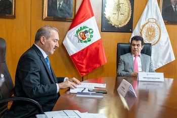 Dos hombres en traje sentados ante una mesa de madera con documentos, frente a banderas y emblemas oficiales de Perú