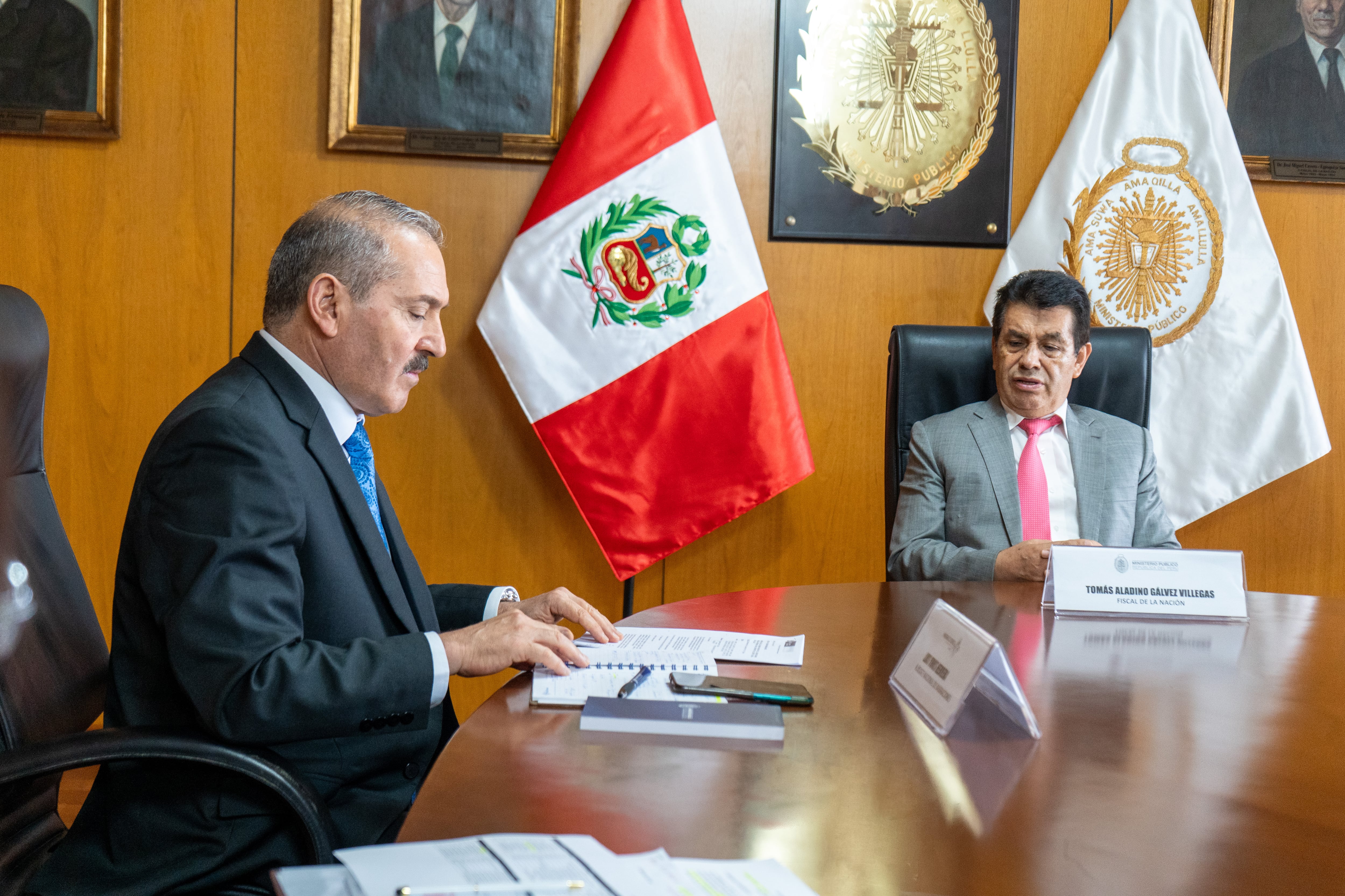 Dos hombres en traje sentados ante una mesa de madera con documentos, frente a banderas y emblemas oficiales de Perú