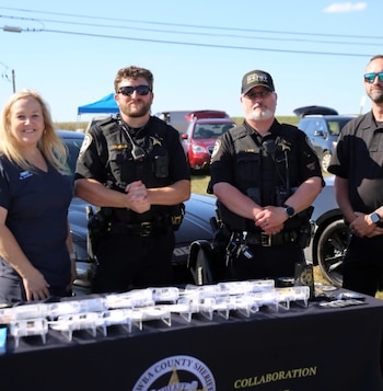Tres agentes del sheriff y una mujer detrás de una mesa con el logo del Sheriff del Condado de Catawba y objetos exhibidos. Vehículos al fondo bajo cielo azul