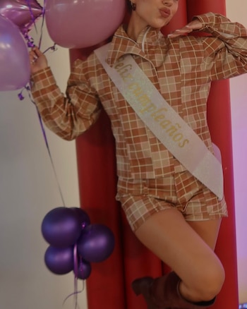 Cami Mayan sonríe sosteniendo un ramo de flores envuelto en papel blanco, vestida con top oscuro, jeans y cinturón dorado, frente a unas escaleras de madera