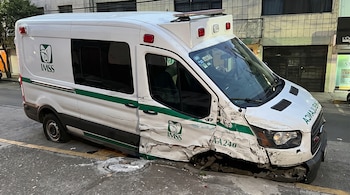 Ambulancia blanca con franjas verdes, abolladuras en el costado delantero derecho y sin la rueda delantera derecha, estacionada en una calle.