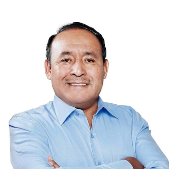 Un hombre con camisa de color azul claro sonríe con los brazos cruzados ante un fondo blanco