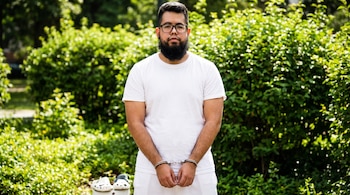 Hombre con barba, gafas, camiseta blanca, short blanco y esposas de pie frente a arbustos verdes. Zuecos blancos se ven en el suelo a su izquierda.