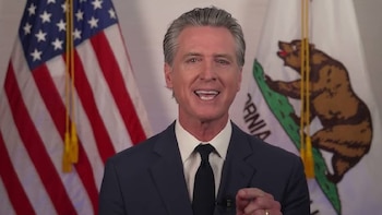Gobernador Gavin Newsom sonriendo, con cabello gris, vestido con traje azul marino y corbata negra. Detrás, la bandera de Estados Unidos y la bandera de California