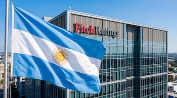 Bandera de Argentina azul y blanca con el Sol de Mayo ondeando frente al moderno edificio de oficinas de Fitch Ratings, con un cielo azul claro de fondo.