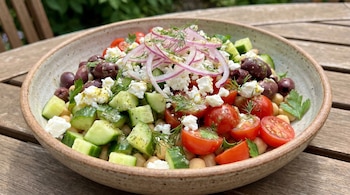 Tazón con ensalada de garbanzos, tomates cherry, pepino en cubos, aceitunas negras, queso feta, cebolla morada en rodajas y eneldo, sobre mesa de madera.