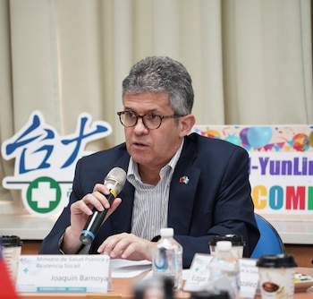 Hombre con gafas, traje, y pin de bandera de Guatemala, hablando por un micrófono. En la mesa, placa con 'Joaquin Barnoya', botellas de agua, vasos de café. Fondo con texto chino