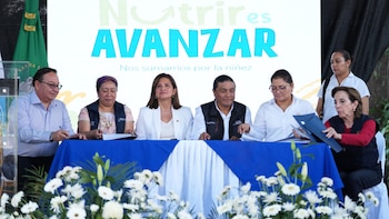 Siete personas, incluyendo funcionarios, sentadas en una mesa larga con mantel azul y flores blancas, frente a un cartel "Nutrir es Avanzar"