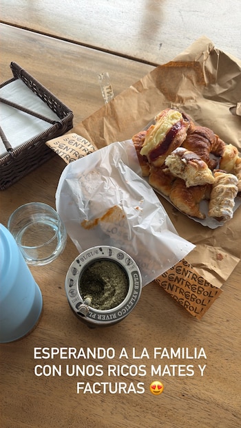 Vista aérea de una mesa de madera con un mate con yerba, un termo azul, un vaso de agua, facturas variadas y un servilletero
