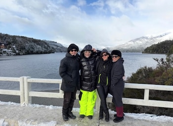 Cuatro personas con ropa de invierno y gafas de sol posan sobre un puente cubierto de nieve, con un lago y montañas nevadas al fondo