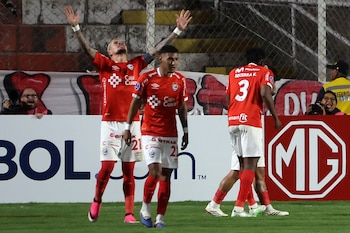 Tres jugadores de fútbol con camiseta roja celebran en el campo de juego, uno con los brazos levantados. Fondo con gradas, público y vallas publicitarias
