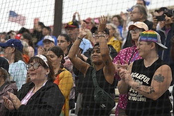 Multitud de personas con vestimenta casual aplauden en un evento al aire libre, vistas a través de una red protectora. Bandera de Estados Unidos al fondo