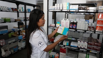 Mujer con uniforme blanco examinando una caja de Amoxicilina y Ácido Clavulánico en una farmacia con estantes metálicos repletos de medicamentos en cajas y frascos