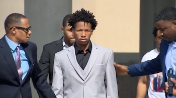 Joven afroamericano con traje gris y cabello rizado oscuro es escoltado por hombres con traje oscuro en un exterior iluminado por el sol