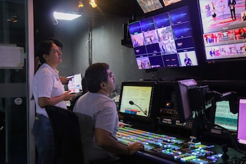 Dos personas en un centro de control de televisión; una mujer de pie y un hombre sentado operando una consola con botones de colores, frente a múltiples pantallas que muestran transmisiones