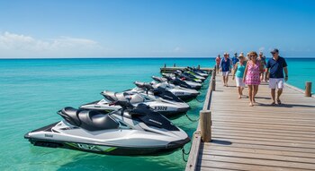 Varias motos de agua blancas y negras están alineadas a lo largo de un muelle de madera sobre el mar Caribe de agua turquesa, con turistas caminando.