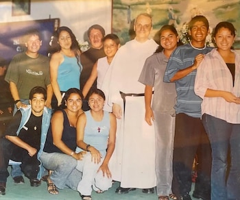 Un religioso vestido con hábito blanco y cordón negro posa de pie en un interior rodeado por nueve personas jóvenes
