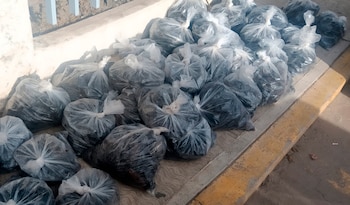 Numerosas bolsas de plástico transparente llenas de un material oscuro apiladas en el suelo de concreto junto a un bordillo amarillo