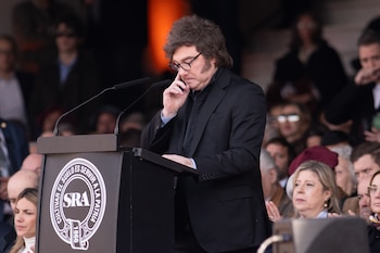 Un hombre de cabello castaño y gafas toca su rostro en un atril con micrófonos y un logo circular blanco de la SRA. Detrás, personas desenfocadas