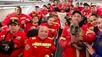 Un grupo de rescatistas con uniformes rojos y chalecos de seguridad, junto a un perro de búsqueda, posa dentro de la cabina de un avión