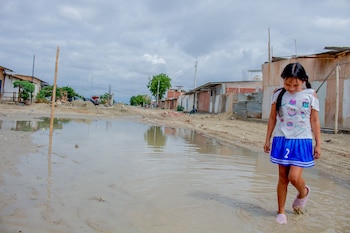 Una niña camina sobre un gran charco de agua en una calle de tierra con viviendas precarias bajo el cielo nublado