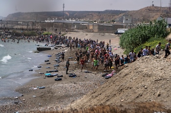 Playa rocosa con cientos de personas corriendo hacia la arena, el mar a la izquierda, neumáticos en la orilla, una valla de seguridad y colinas al fondo