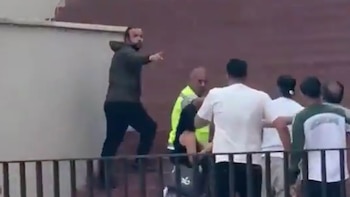 Un hombre en sudadera oscura de pie sobre una cornisa señala hacia un grupo de personas, una de ellas con un chaleco de seguridad amarillo