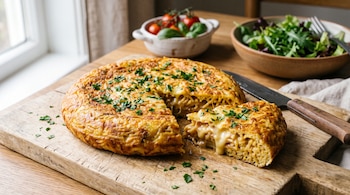 Tortilla de fideos cortada con queso derretido en una tabla de madera, acompañada por un cuchillo, tomates cherry y un tazón de ensalada.