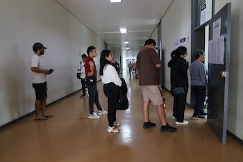 Varias personas están de pie en un pasillo largo y bien iluminado de un edificio, algunas mirando papeles pegados en la puerta de un aula marcada "511"