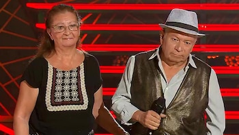 Un hombre y una mujer de pie en un escenario con luces rojas de fondo. El hombre lleva sombrero, chaleco y camisa, y sostiene un micrófono