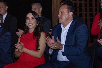 Un hombre de traje azul y camisa blanca y una mujer con vestido rojo aplauden sentados junto a otras personas